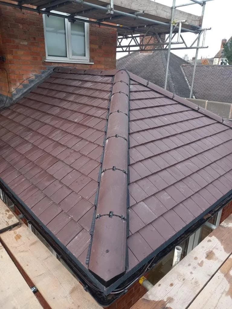 The finished roof over the rear extension: new red-brown interlocking tiles laid up to a dry ridge system with black union clips, and a dormer window below the scaffold.
