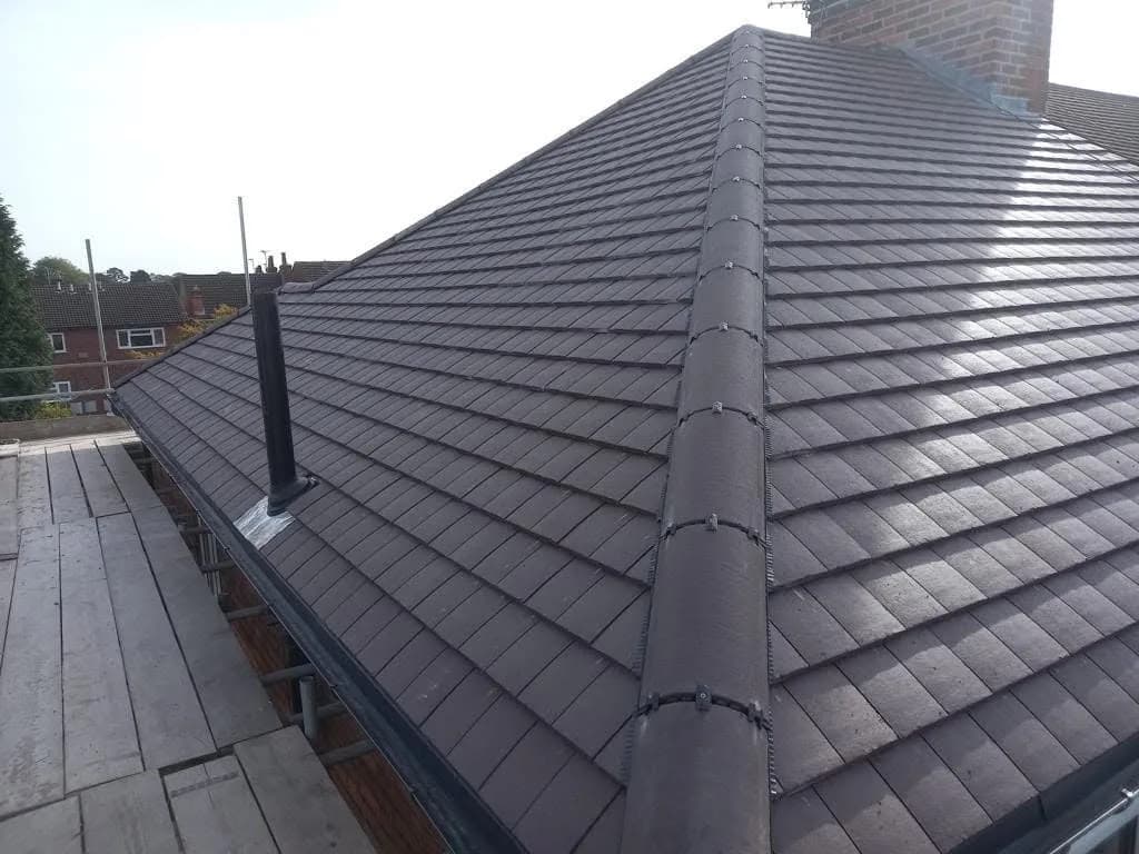 The finished main roof slope: new red-brown tiles running up to a dry ridge system, a black soil vent pipe dressed with new flashing near the top, and rooftops of neighbouring houses beyond.