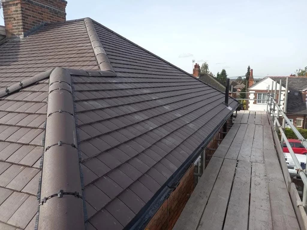 The finished hip and dry ridge on the main roof, seen along the scaffold boards towards the street, with a neighbouring property and a parked car below under a clear blue sky.