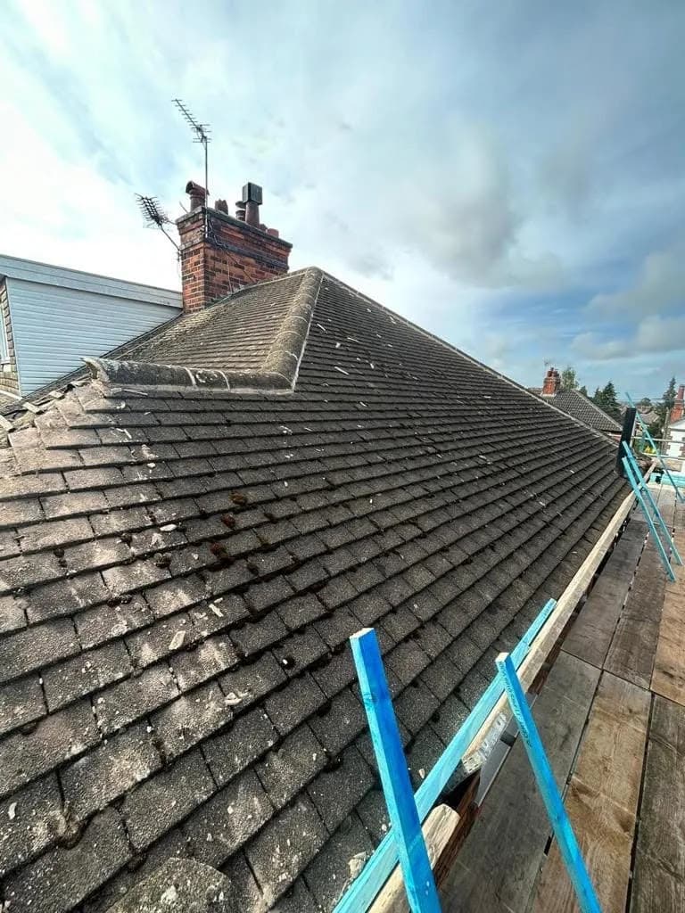 The roof before work: weathered grey concrete interlocking tiles across a hipped slope, a brick chimney stack with a television aerial, and scaffold boards along the eaves under a cloudy sky.
