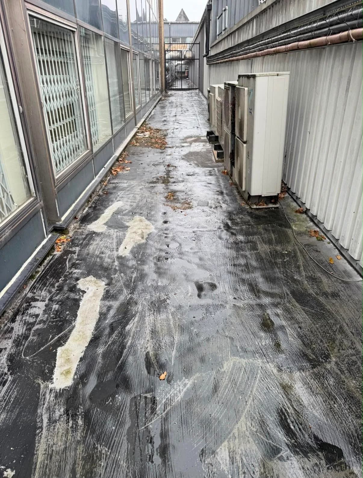 The flat roof walkway between two factory buildings before the overlay, its surface streaked with old repair compound and scattered debris, with a row of air-conditioning units along one side.