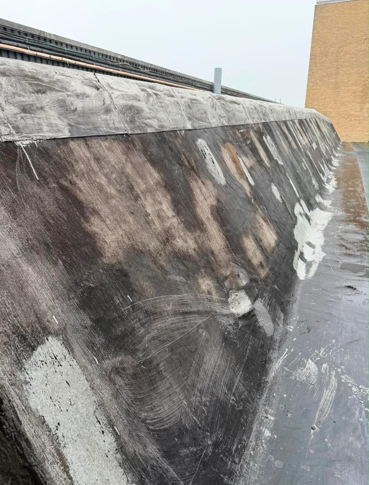 Close view along a worn slope of the factory roof, the felt smeared and patched from previous repair attempts, beside a rain-soaked valley.