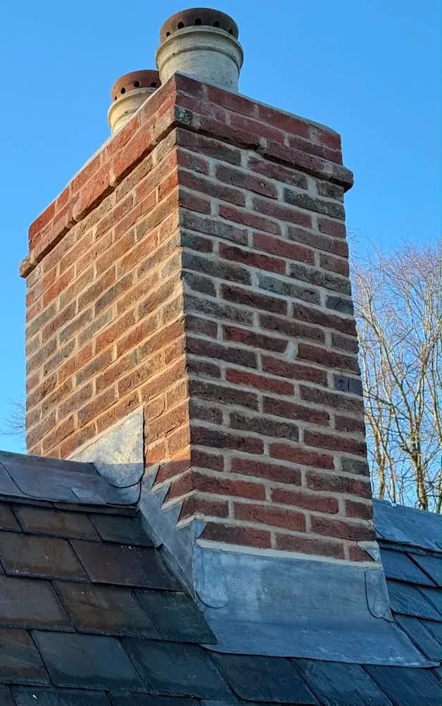 The same chimney stack after the work, its brickwork repointed in pale lime mortar with fresh flaunching around the pots and new lead dressed neatly where the stack meets the finished slate roof.