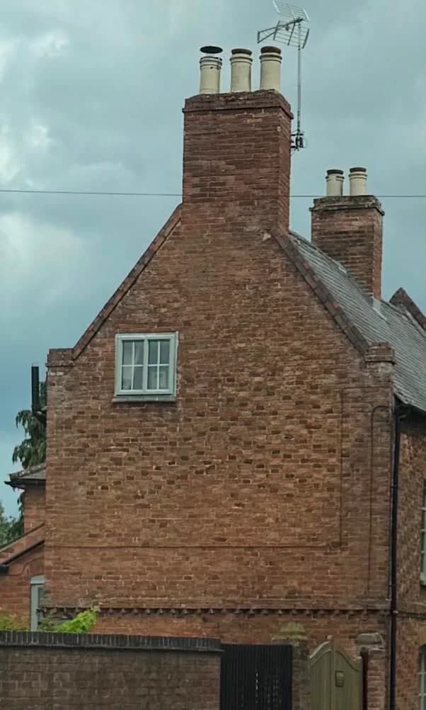 The gable end of the house before work, seen from the road under a grey sky: worn red brickwork with patchy, weathered pointing, a tall chimney stack with three pots and a television aerial, and a single small casement window.
