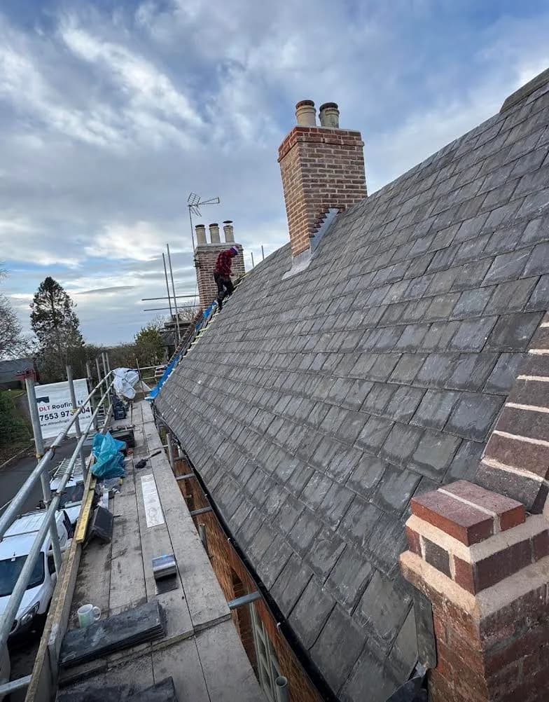 The re-slating part way through, seen along the roof from the scaffold: new blue-grey slate laid up most of the slope with a roofer working near the far chimney, the repointed stacks finished and the apex still open before the lead ridge went on.