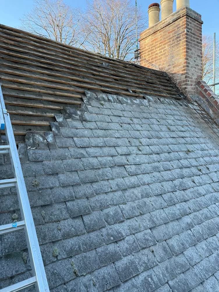 Stripping underway: the top third of the roof slope opened up to bare timber battens and rafters while the lower two thirds still carry the old grey slates, with a ladder resting against the slope and the brick chimney alongside.