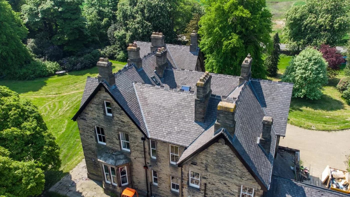 The completed restoration in sunlight — the full natural-slate roof relaid across the period stone house, its original stone chimney stacks kept in place, framed by mature trees and lawns.