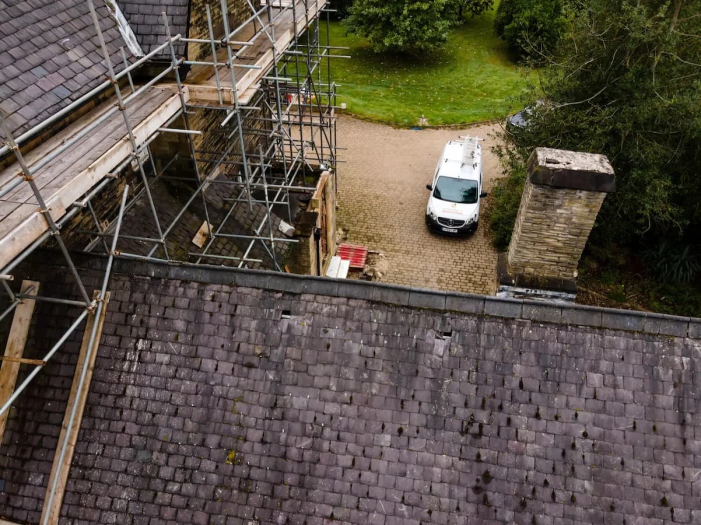 Scaffolding being raised against the stone gable at the start of the job, with the aged, moss-flecked slate roof still in place in the foreground and a DLT Roofing van parked on the driveway below.