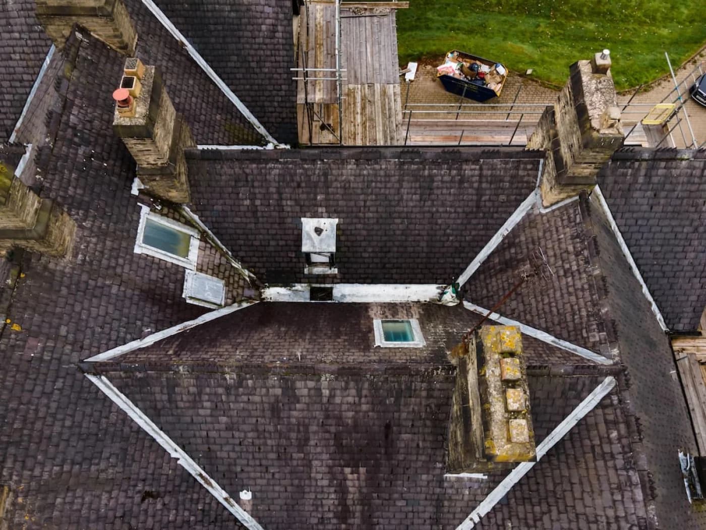 Overhead drone view of the property's original natural-slate roof before restoration — weathered, moss-darkened slates with worn lead flashing running along the valleys and around the stone chimney stacks, and a skip on the terrace below.