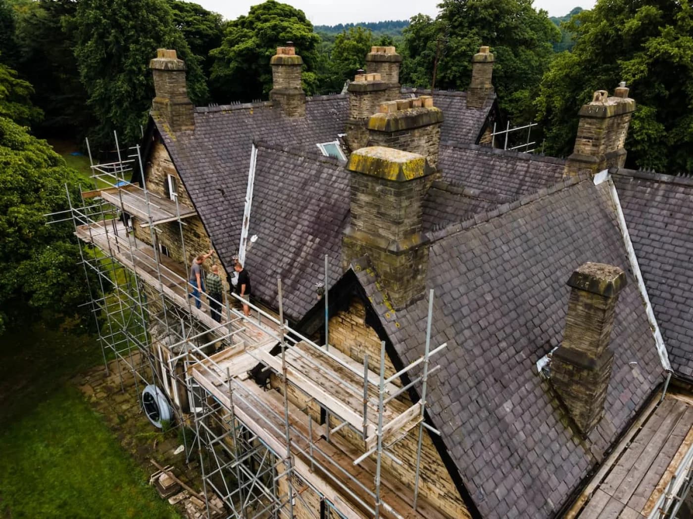 The period stone house fully scaffolded mid-project, with three roofers working along the boards beneath a run of tall stone chimney stacks and the slate roof partly stripped back.