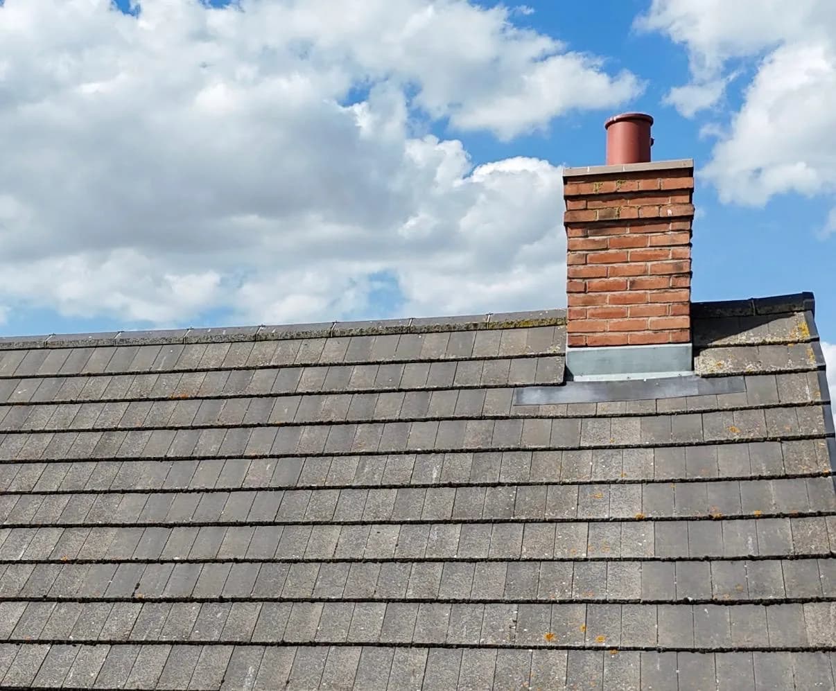 Close-up of a red-brick chimney stack with a dark red pot, grey GRP flashing at its base and lead soakers, set into a grey concrete-tiled roof slope.