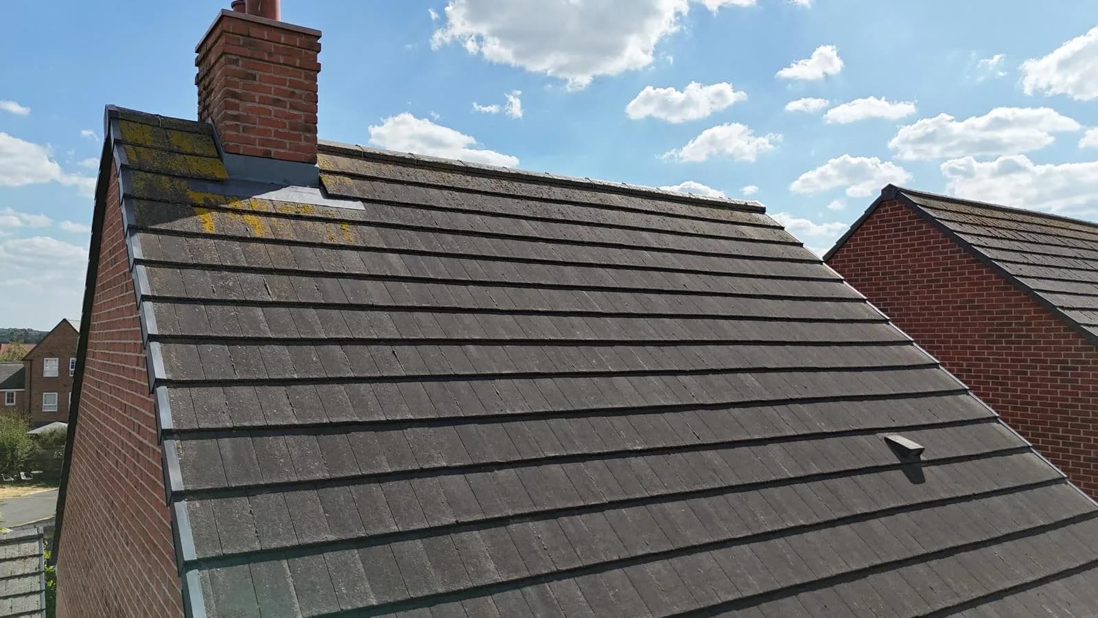 A wide drone view across a grey concrete-tiled roof slope, with a patch of yellow-green lichen near the chimney and neighbouring rooftops visible in the background.