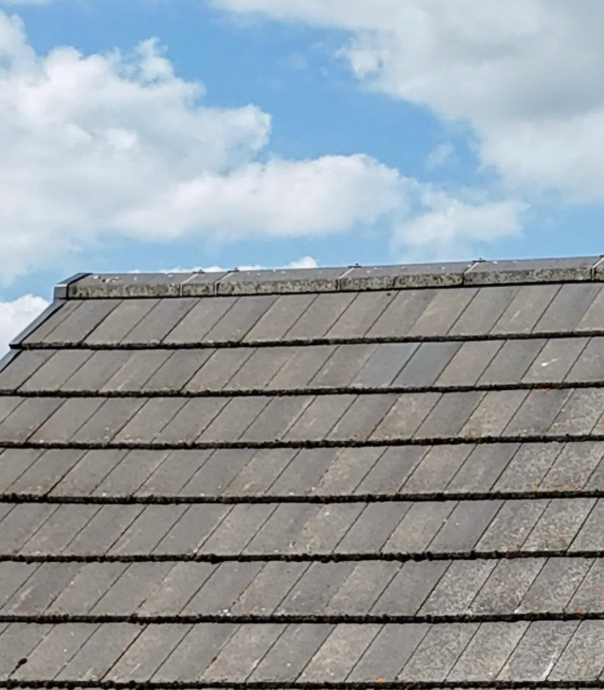 A drone's-eye view along a dry ridge line on a grey concrete-tiled pitched roof, with the capped ridge tiles and gable verge meeting under a partly clouded sky.