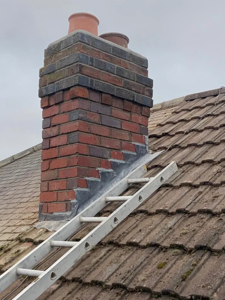 A red-brick chimney stack with two clay pots and freshly fitted lead stepped flashing and back gutter, a ladder resting on the tiled roof.