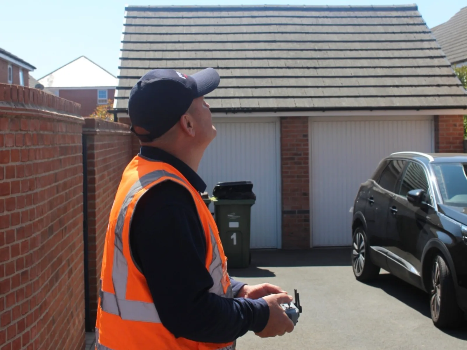 Dave from DLT Roofing, in an orange hi-vis vest and navy cap, standing on a driveway with a drone controller in both hands, looking up at the moss-flecked concrete-tiled roof of a double garage under clear blue sky.