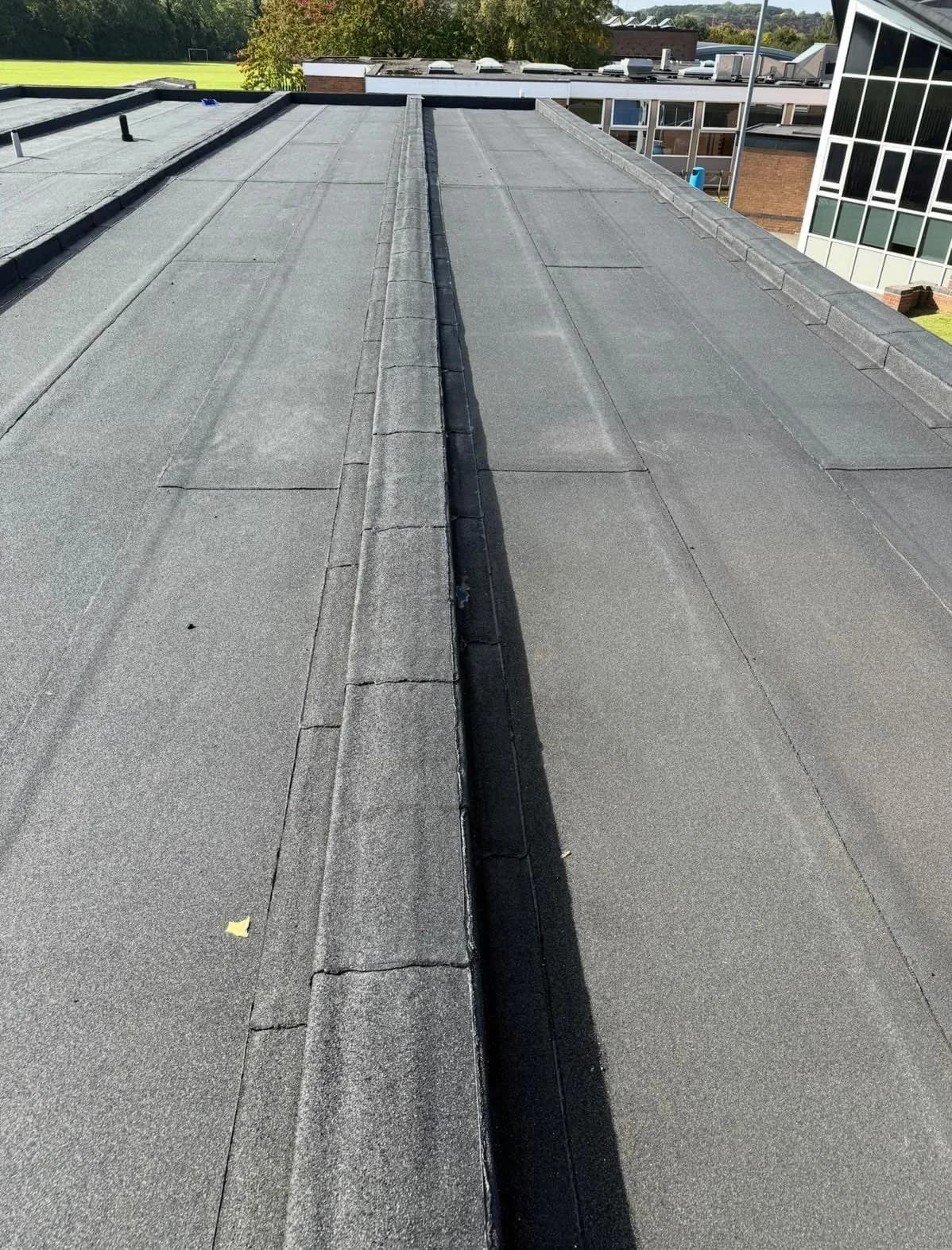 The same roof from the same vantage point after renewal, in sunshine — a continuous dark grey mineral-surfaced bitumen membrane laid across both bays, with neatly lapped seams and the central gutter dressed the full length of the run.
