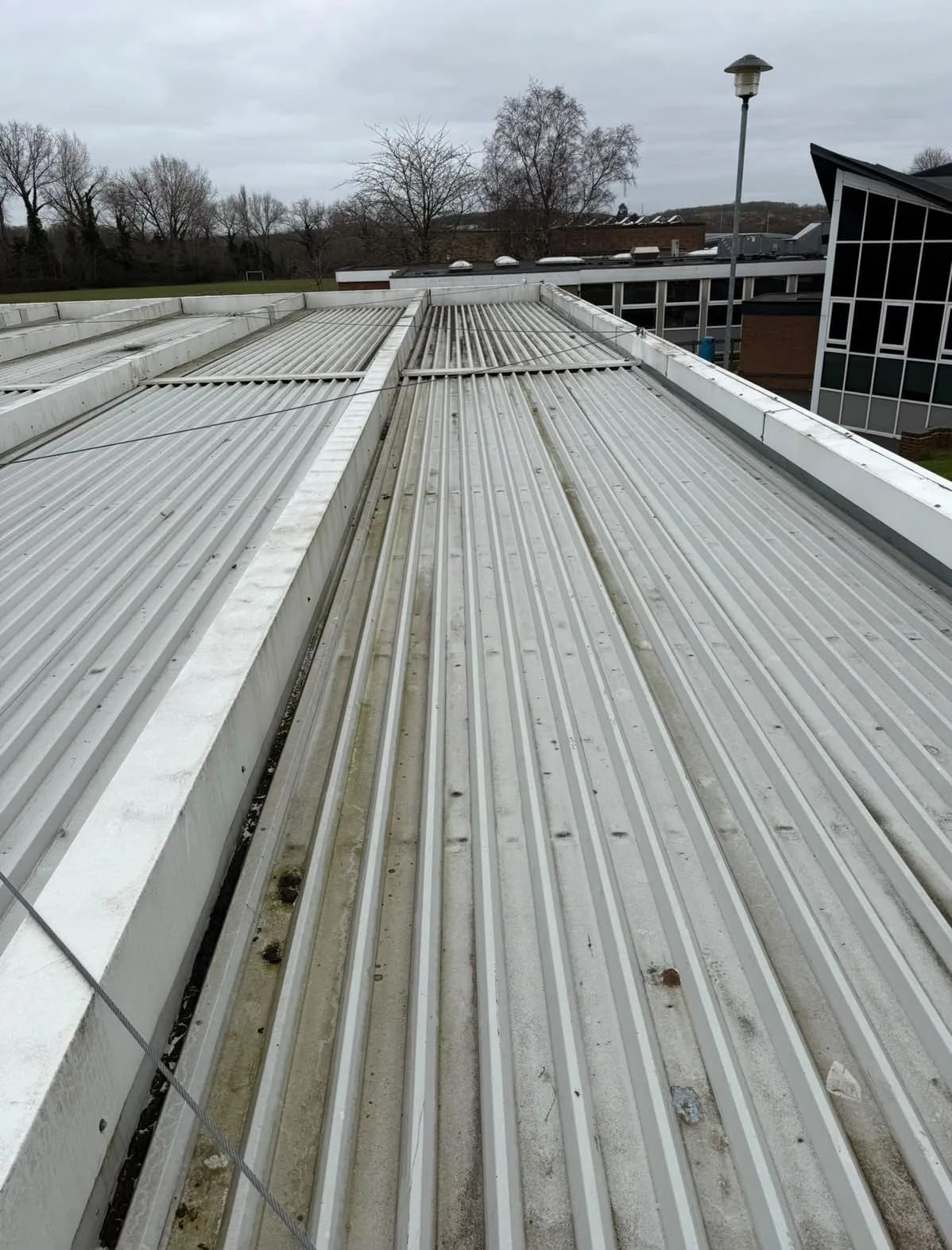 The original profiled metal sheet roof before renewal, under flat overcast light — long runs of weathered grey-white cladding either side of a central gutter channel, with dirt and moss staining collected along the seams and gutter.