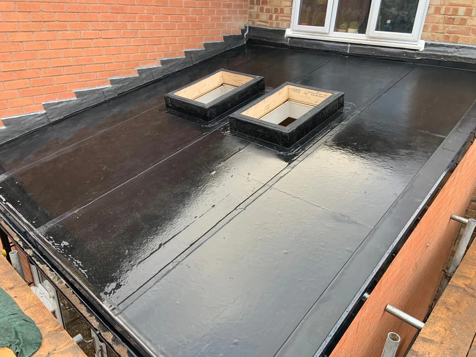 A glossy black flat roof with two timber-framed rooflight upstand kerbs and stepped lead flashing against a red-brick wall.