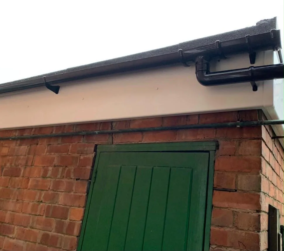 Close-up of new black guttering and a downpipe hopper fixed to a white fascia and soffit board on a brick outbuilding, above a green timber door.