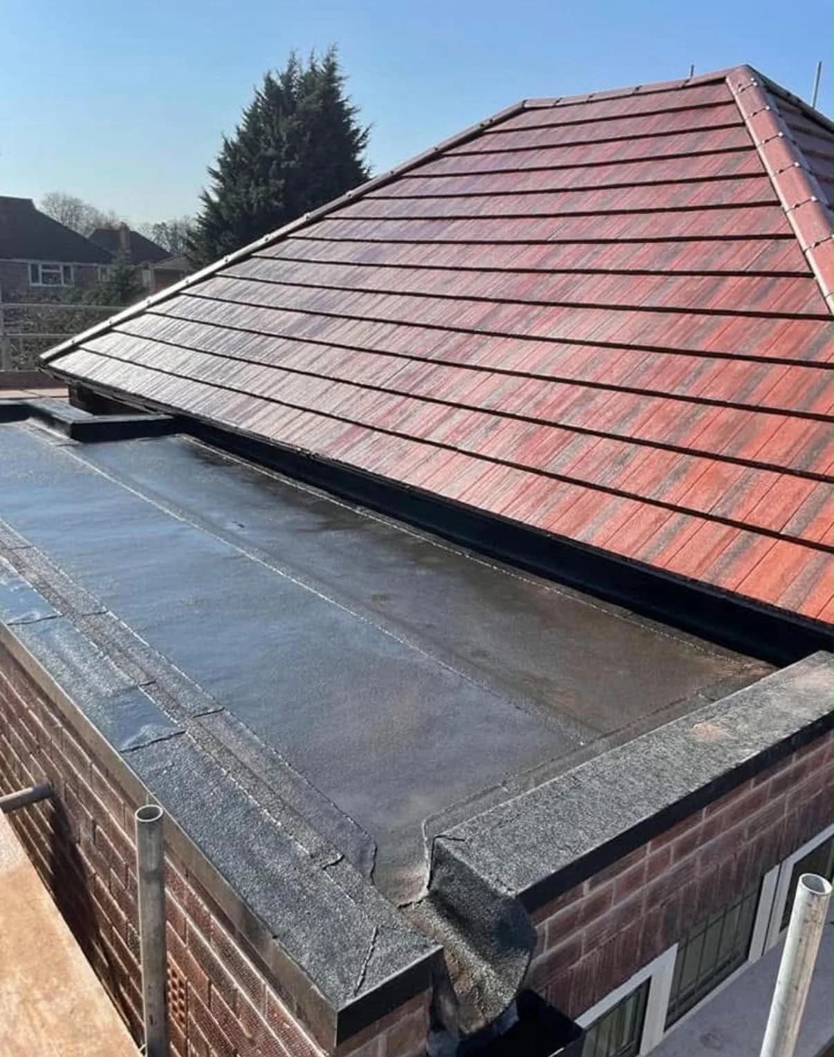 The same roofline from the same vantage point after renewal, in sunshine: fresh red clay tiles on the pitched roof above, a clean new eaves and gutter line, and a smooth, newly laid flat roof with neat welted corners below.