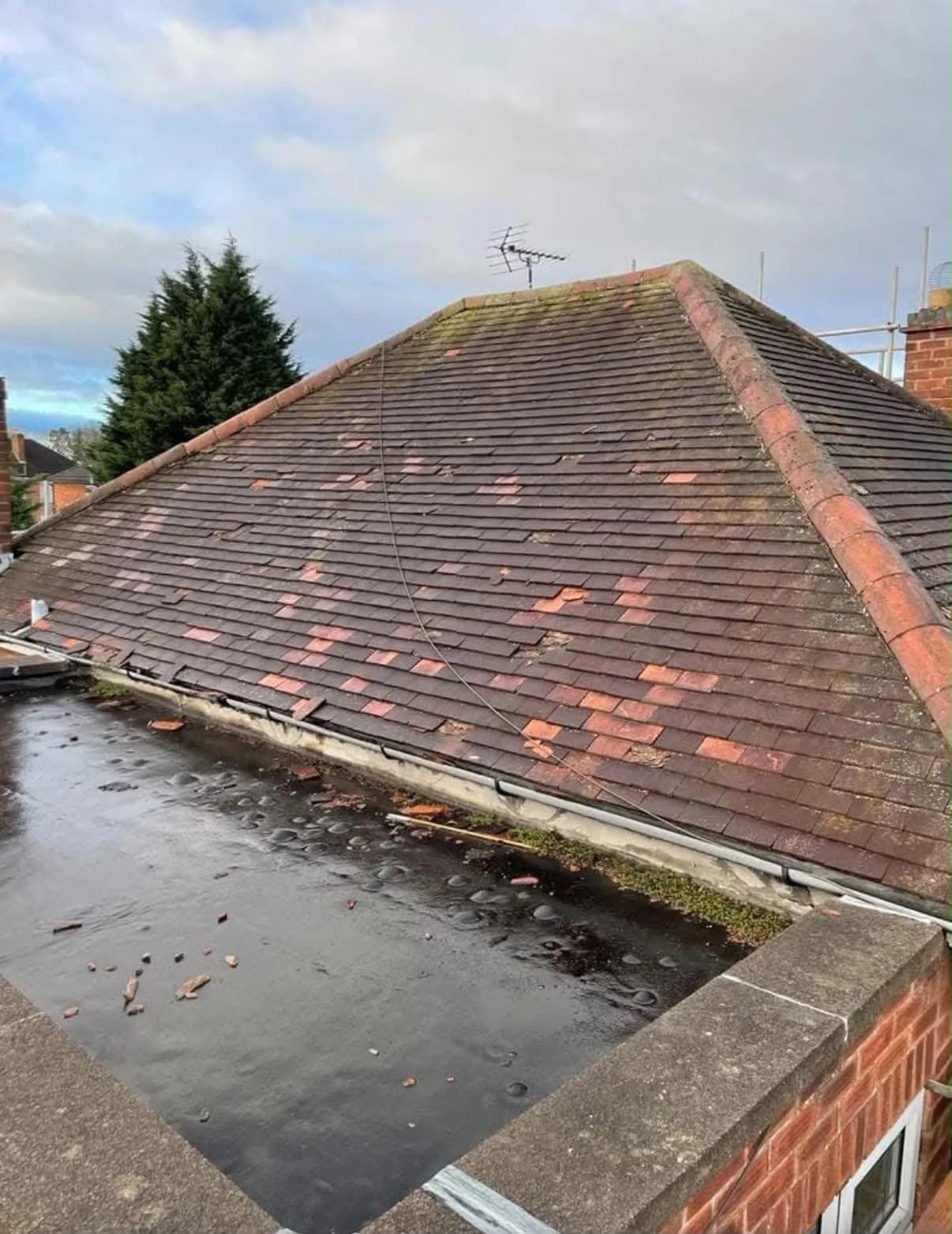 The original roofline before renewal, seen from the flat roof below: cracked and slipped red clay tiles on the pitched roof above, a perished, gapped gutter line along the eaves, and a flat roof surface littered with broken tile fragments and standing water.