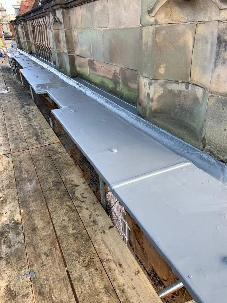 A long parapet gutter behind a carved stone balustrade on a heritage building, freshly waterproofed with a grey membrane, timber walkway boards laid alongside.
