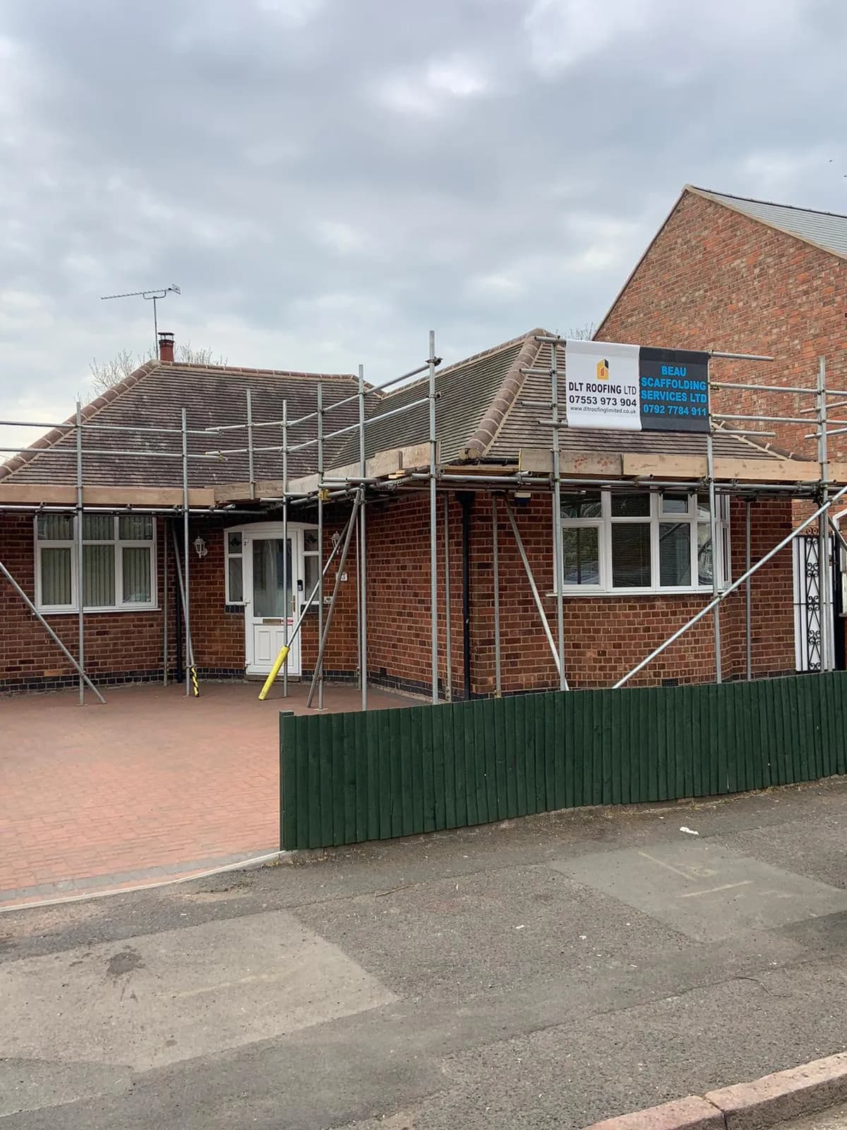 A bungalow being re-roofed behind scaffolding, with a DLT Roofing banner fixed to the scaffold and the new hip roof partly tiled.