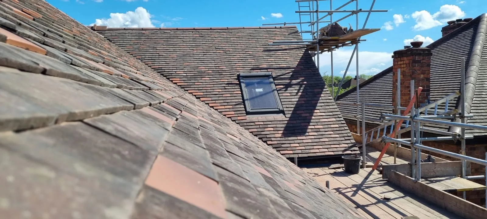 An older clay plain-tile roof being stripped and re-laid, with a roof window fitted and scaffolding and chimney stacks behind under a blue sky.