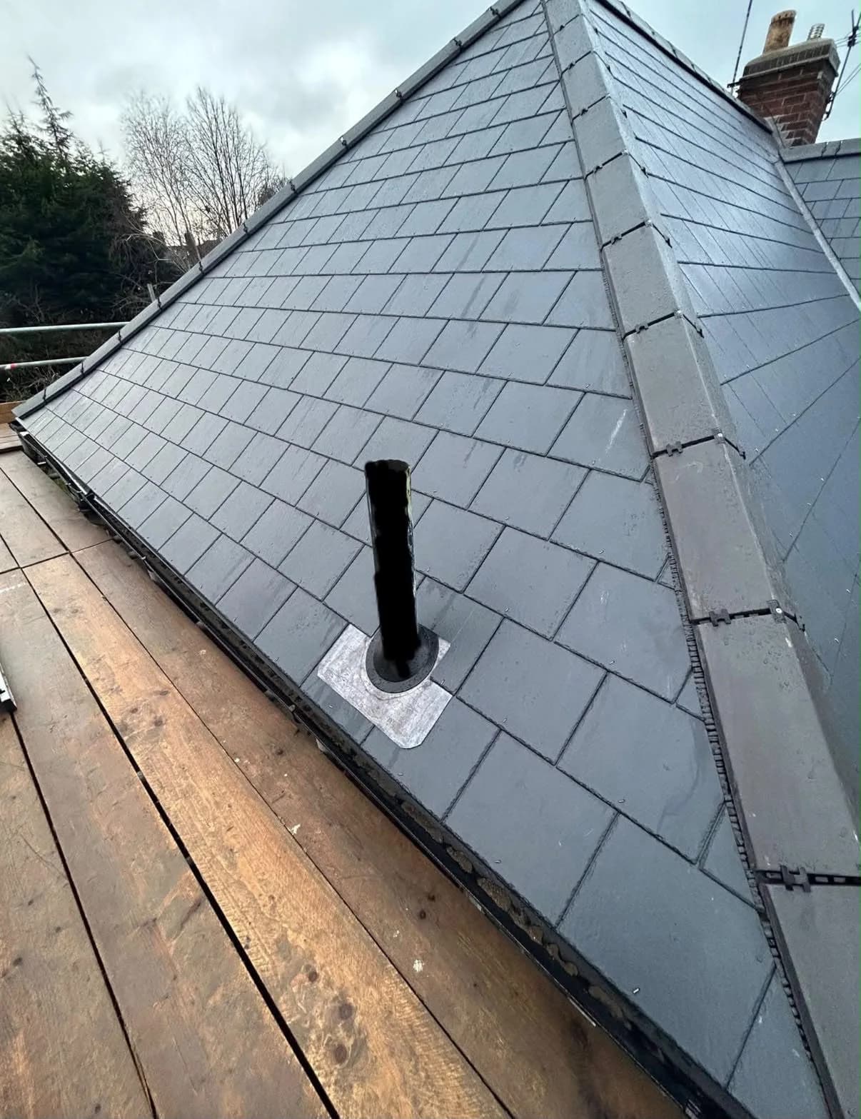 A close, roof-level view of a newly re-slated pitched roof in dark grey slate: neat overlapping rows of new slate running up to a capped hip, a soil-vent pipe with a lead flashing collar, and a wooden scaffold board in the foreground.