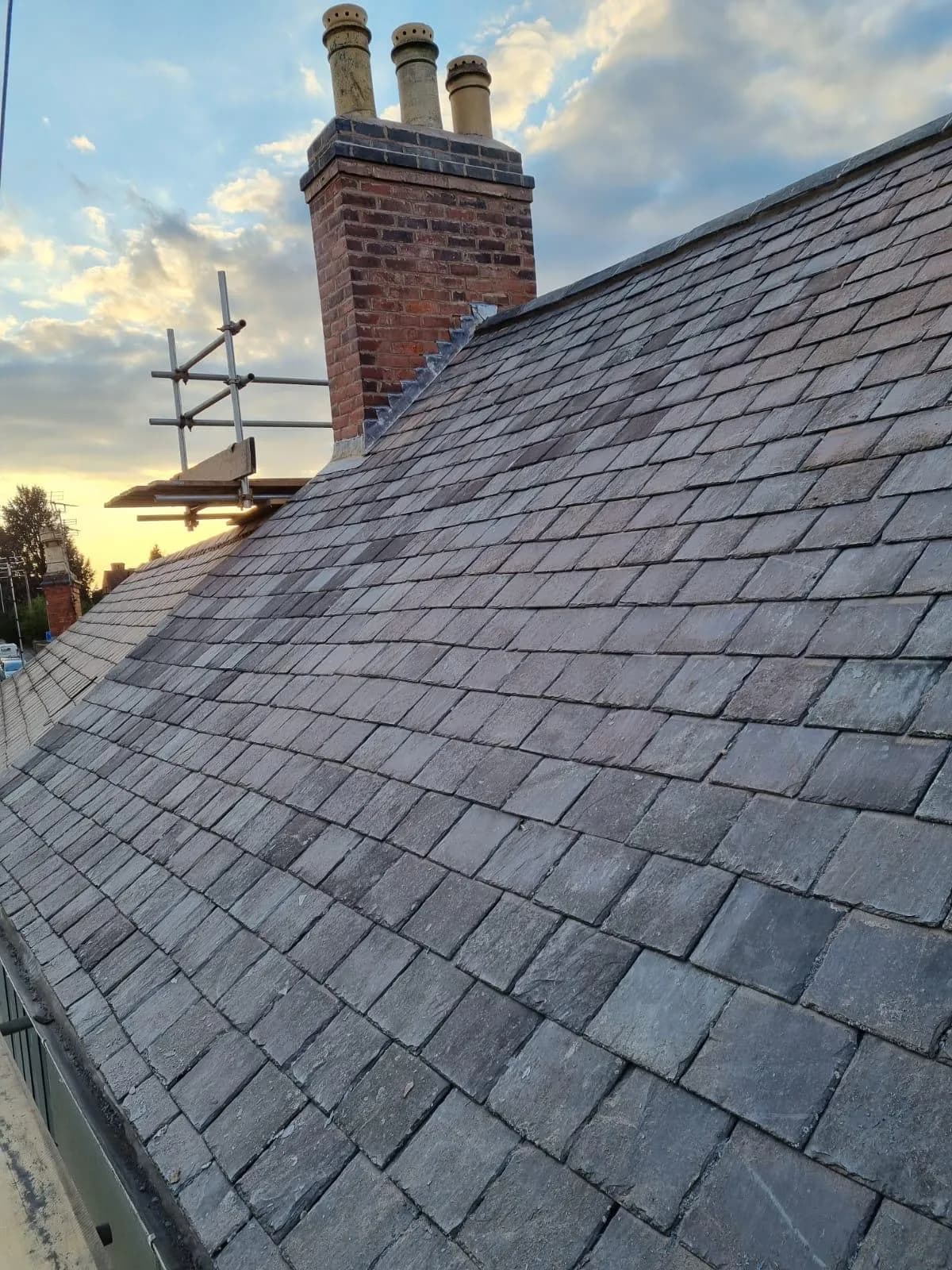 A newly laid natural-slate pitched roof meeting a tall red-brick chimney with clay pots, photographed at dusk with scaffolding to one side.