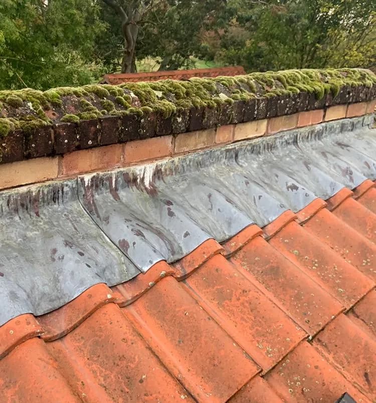 A weathered roof verge with moss-covered ridge tiles and old, discoloured lead soakers against clay pantiles, before the verge was rebuilt.